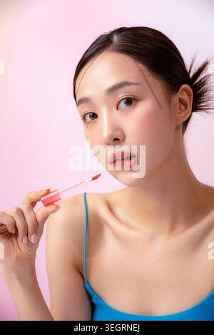 A young Asian woman is applying lip gloss in a studio setting. Stock Photo