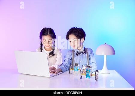 Two children sitting at a table, using a laptop and playing with a robotic car. Stock Photo