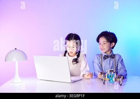 Two children, one boy and one girl, sit at a desk together, both using a laptop to build a robot. Stock Photo