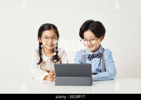 Two children in glasses sit at a table, using a tablet. Stock Photo