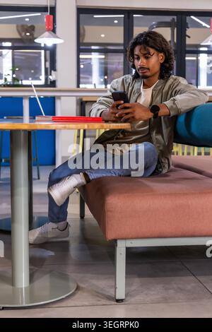 Adult Indian man in olive jacket sitting on cushioned bench in cafe using smartphone, notebook, cup Stock Photo