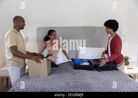 Diverse family packing clothes and lifting lamp on gray quilted bed with open suitcase Stock Photo