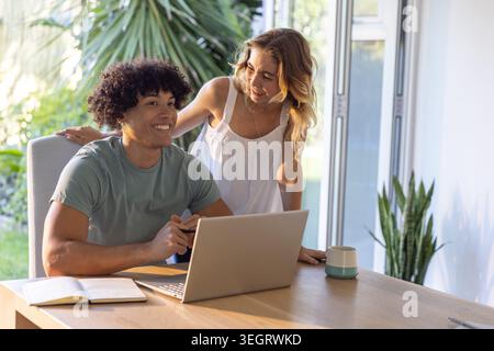 Diverse couple leaning, working at home table, man in green T-shirt, woman white dress using laptop Stock Photo