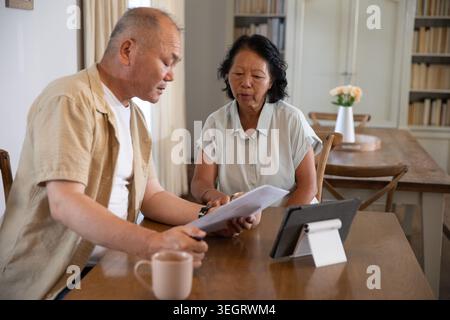 Asian couple examining printed documents at home dining table while using tablet and coffee mug Stock Photo