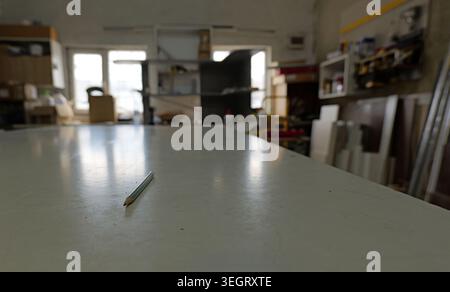 Pencil lying on a large white workbench surface in a woodworking or carpentry workshop, with blurred tools, materials, and a window visible in the bac Stock Photo