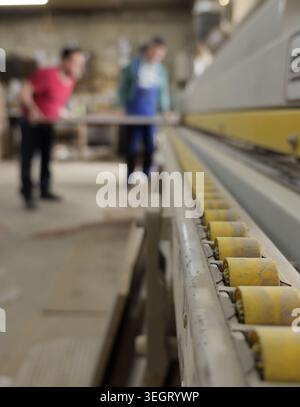 Woodworkers are operating an industrial edge banding machine, creating furniture components in a busy workshop setting, showing part of the production Stock Photo