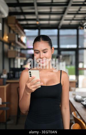 A vertical, authentic, unretouched photo of a happy young Thai woman from the Isan region in her 20s, smiling while using phone inside a modern coffee Stock Photo