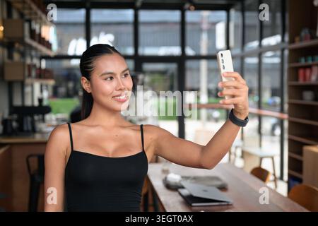 An authentic, unretouched horizontal photo of a happy young Thai woman from the Isan region in her 20s, smiling while holding up her smartphone to tak Stock Photo