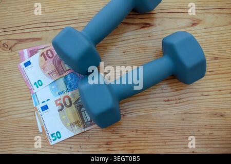 High angle shot of fifty and ten Euro banknotes placed under heavy dumbbells on a wooden table. Conceptual representation of the high cost of fitness Stock Photo