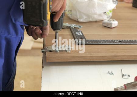Professional handyman fixing metal hinges to a wooden panel using an electric power drill and screws, working in a workshop or home renovation project Stock Photo