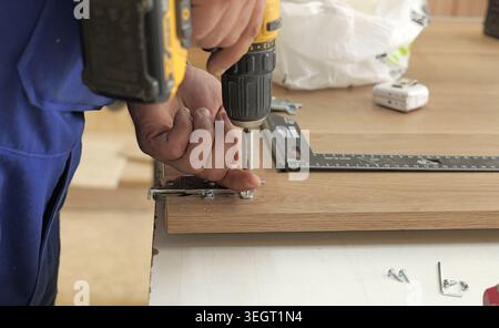 Worker using an electric drill to fasten screws into a wooden board, building furniture or cabinetry in a workshop environment, focusing on constructi Stock Photo