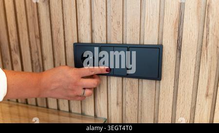 Close up of woman pressing modern black light switch on wooden wall panel with electrical socket in contemporary interior. Stock Photo
