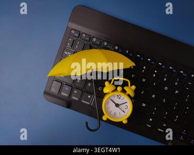 A small yellow umbrella shields a miniature yellow alarm clock sitting on top of a black computer keyboard. Stock Photo