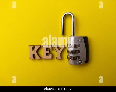 An open silver combination padlock rests next to the wooden letters spelling "KEY" on a bright yellow background. Stock Photo