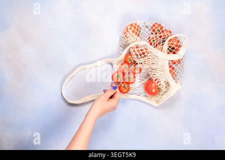 Hand holding a bunch of fresh, red cherry tomatoes still on the vine, pulling them from a reusable cotton mesh bag filled with more tomatoes, promotin Stock Photo