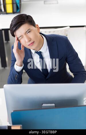 A young businessman in a suit sits at his desk, rubbing his temples in frustration, staring at his computer screen. Stock Photo