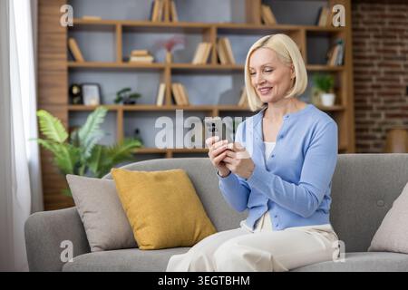 Blonde woman sitting comfortably on a sofa, smiling while interacting with a smartphone, representing modern communication and digital lifestyle in a cozy home environment Stock Photo