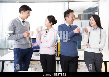 Four colleagues are standing in an office, chatting and holding cups of coffee. Stock Photo