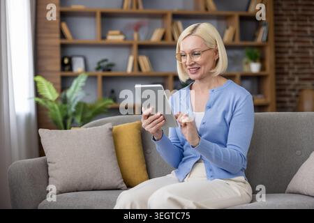 Senior woman in glasses and a blue cardigan smiling on a living room sofa while tapping a digital tablet, relaxed and engaged in browsing, reading or video calling Stock Photo