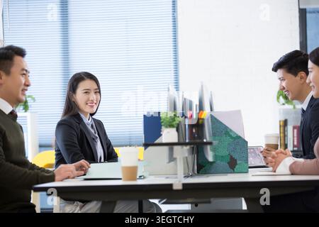 A group of colleagues are laughing and chatting around a table in a modern office. Stock Photo
