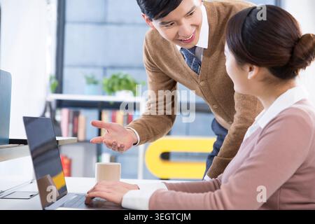 A young Asian male colleague explaining something to a young Asian female colleague at a desk in an office. Stock Photo
