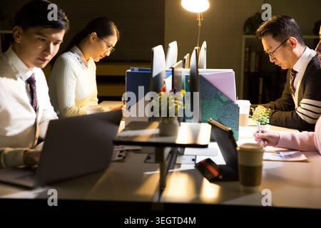 Colleagues working late at night in a dimly lit office, focused on their laptops. Stock Photo