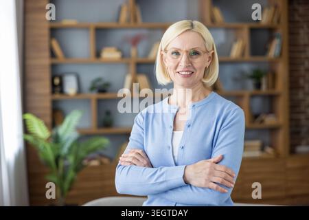 Confident blonde woman with glasses smiling and arms crossed in front of a modern wooden bookshelf, projecting professionalism, expertise and a positive business or education vibe Stock Photo