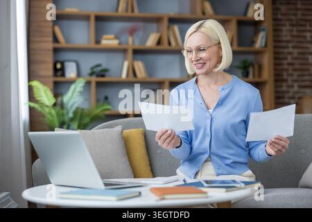 Mature businesswoman in cozy home office reviewing documents and checking laptop for finance and online work, smiling confidently while organizing papers at her desk Stock Photo