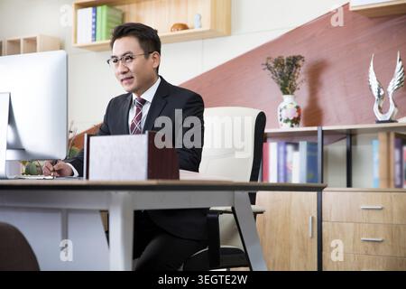 A young Asian businessman sits at his desk, focused on his computer screen in a modern office. Stock Photo