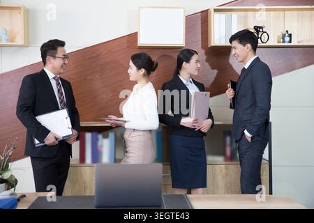 A group of diverse business professionals in formal attire stand in a modern office, engaged in a discussion. Stock Photo