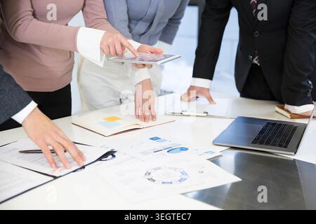Business professionals gathering around a white table, examining papers and a tablet, in a professional office setting. Stock Photo