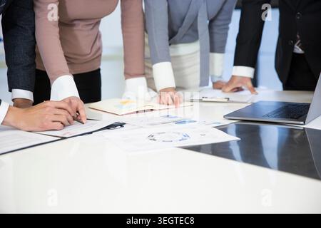 A group of businesspeople gather around a table, examining documents and laptops. Stock Photo