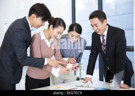 A group of four Asian colleagues in business attire are gathered around a table, reviewing documents and using a tablet together in a modern office setting. Stock Photo