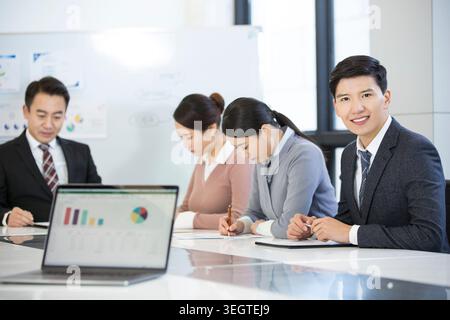 A diverse group of businesspeople sit around a conference table, engaged in a meeting, with a laptop displaying a bar chart on the table in front of them. Stock Photo