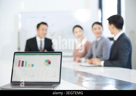 Laptop screen displaying a pie chart and bar graph on a white table in a meeting room with four people in the background. Stock Photo