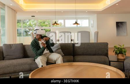 African American man sitting on sofa using VR-headset holding remote by coffee-table in living room Stock Photo