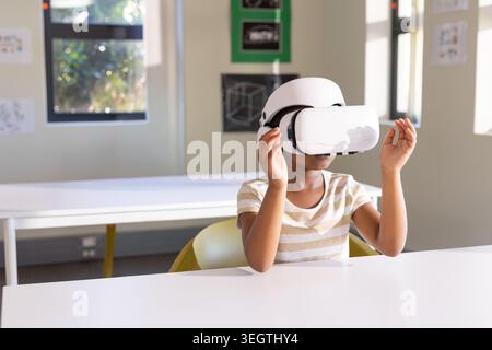 African American child adjusting white virtual reality headset while sitting at classroom table Stock Photo