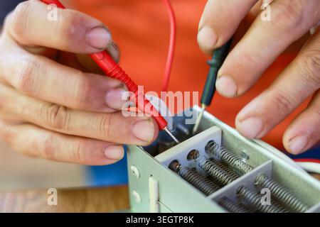 Focused electrician hand testing electrical component with red and black multimeter probe measuring voltage during repair maintenance work in workshop Stock Photo