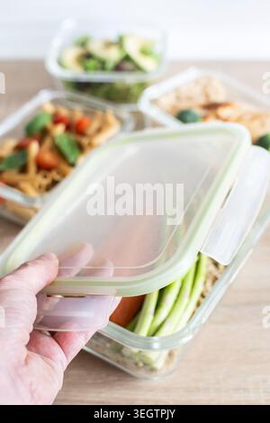 Hand closing a glass meal prep container filled with salmon, quinoa and fresh vegetables on a kitchen table. Healthy eating and weekly food preparatio Stock Photo