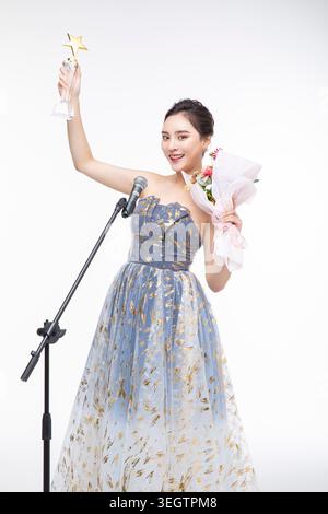 A young woman in a formal dress holds a microphone and raises a trophy in a studio setting. Stock Photo