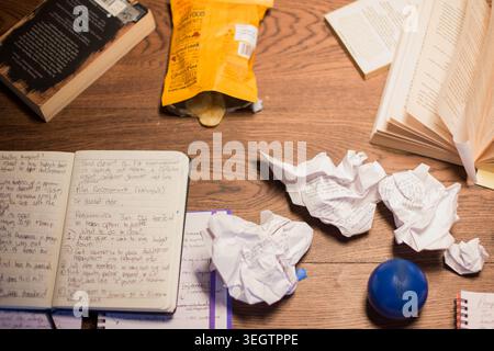 A cluttered desk with an open notebook and screwed up script pages. Snacks, a stress ball, and books sit on the table Stock Photo