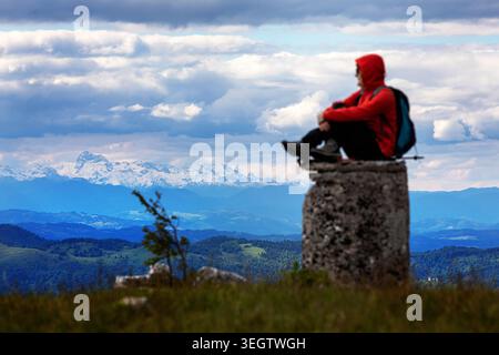 Hiker standing on top of Baba hill on a Wooded Javorniki mountain range with view of a snowy julian alps and highest peak Triglav, slovenia Stock Photo