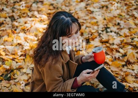 Person sits on ground using smartphone while holding thermal cup in park filled with fallen leaves. Checking banking app, using mobile navigation tool Stock Photo