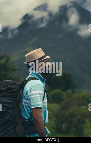 A profile view of a bearded man wearing a hat and backpack looking at the misty mountain landscape at Ali Bedni Bugyal, Chamoli, Uttarakhand, India. Stock Photo