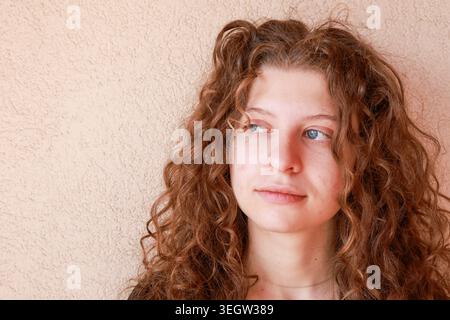 A young woman stands by a light wall with curly hair. She gazes away from the camera with a thoughtful expression. Her hair frames her face, and the l Stock Photo