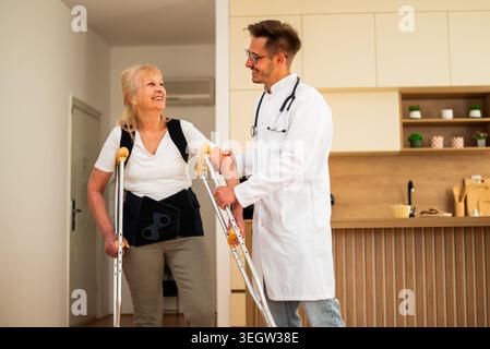 A doctor supports an elderly woman using crutches, helping her walk safely at home during recovery from an injury. Stock Photo