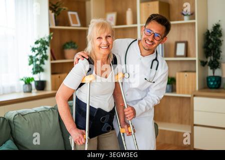 A doctor supports an elderly woman using crutches, helping her walk safely at home during recovery from an injury. Stock Photo