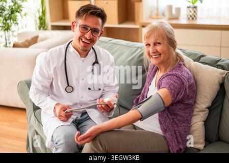 A doctor measures an elderly woman’s blood pressure during a home visit, ensuring regular health monitoring and care. Stock Photo