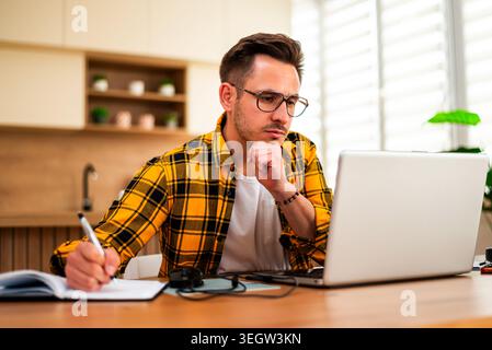 With a pensive expression, a man works on his laptop and writing in note pad, appearing to analyze information or solve a problem online. Stock Photo