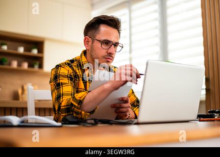 Freelancer in a yellow plaid shirt looks pensive as he reviews a document and data on his laptop. Stock Photo
